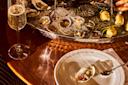 A glass of champagne and a plate with a single oyster sit on a wooden table beside a large ice-filled seafood platter with oysters, clams, and lemon wedges at The Dynamo Room restaurant.
