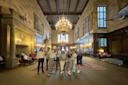 A group of people stand in a large, ornate hall with high ceilings, chandeliers, and wooden paneling. Tapestries hang on the walls, and comfortable chairs are arranged near a carpet. Large windows allow natural light to illuminate the space.