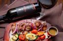 A wooden platter with grilled meat and vegetables, including zucchini, tomatoes, and carrots, garnished with red onion slices. A small cup of sauce is on the side. A bottle of red wine and a filled wine glass accompany the dish.