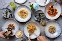 A table with various plated dishes, including seafood and pasta, accompanied by seaweed, sliced apples, and drinks. Hands hold forks, reaching for food. The setting suggests a shared dining experience.