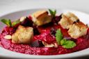 A close-up of a vibrant beetroot dip garnished with croutons, beet chunks, almond slivers, mint leaves, and a drizzle of olive oil, served in a white bowl.