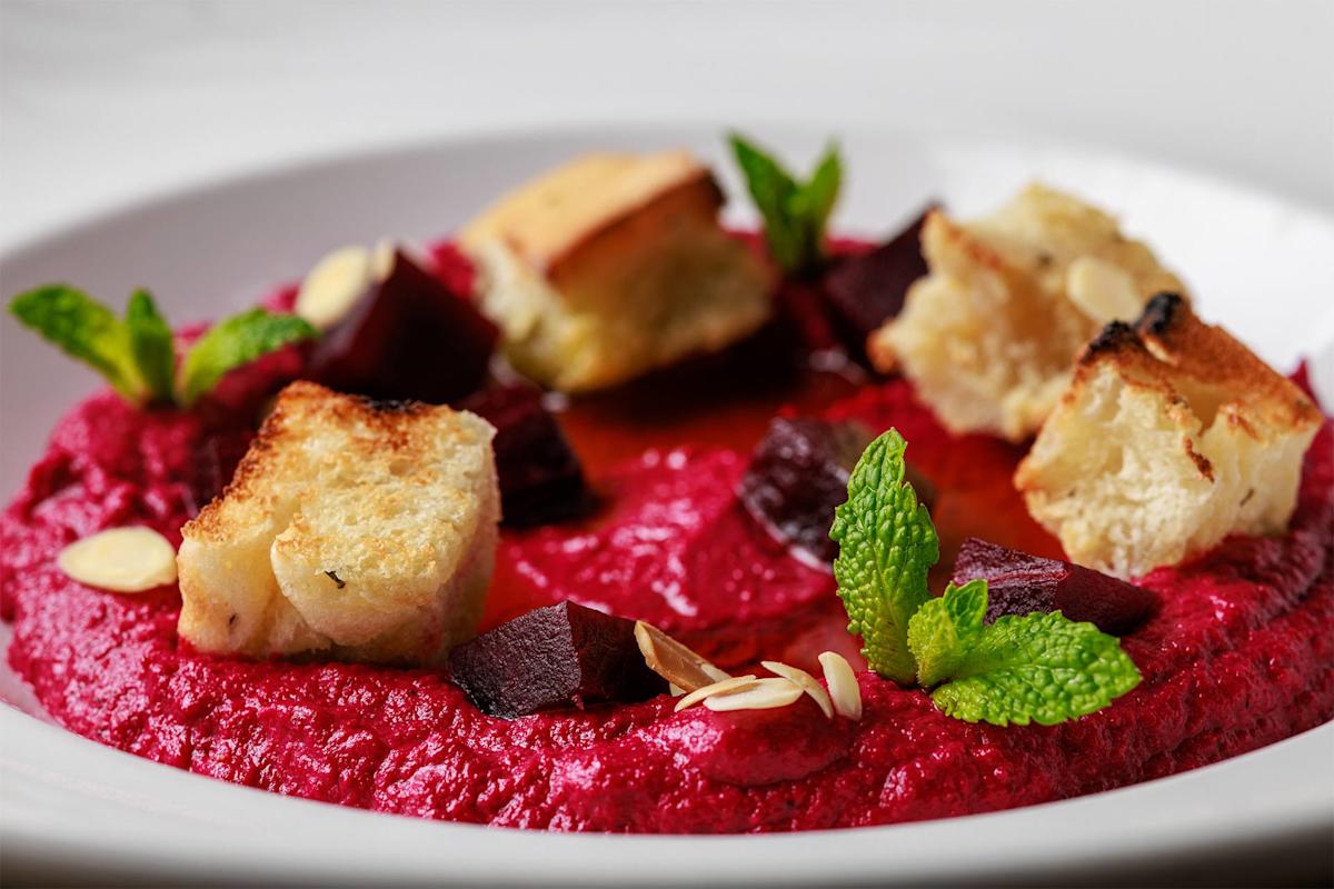 A close-up of a vibrant beetroot dip garnished with croutons, beet chunks, almond slivers, mint leaves, and a drizzle of olive oil, served in a white bowl.