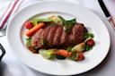 A white plate with sliced roast beef, glazed carrots, baby potatoes, sugar snap peas, and cherry tomatoes, garnished with herbs, resting on a white tablecloth with a fork and knife beside it.