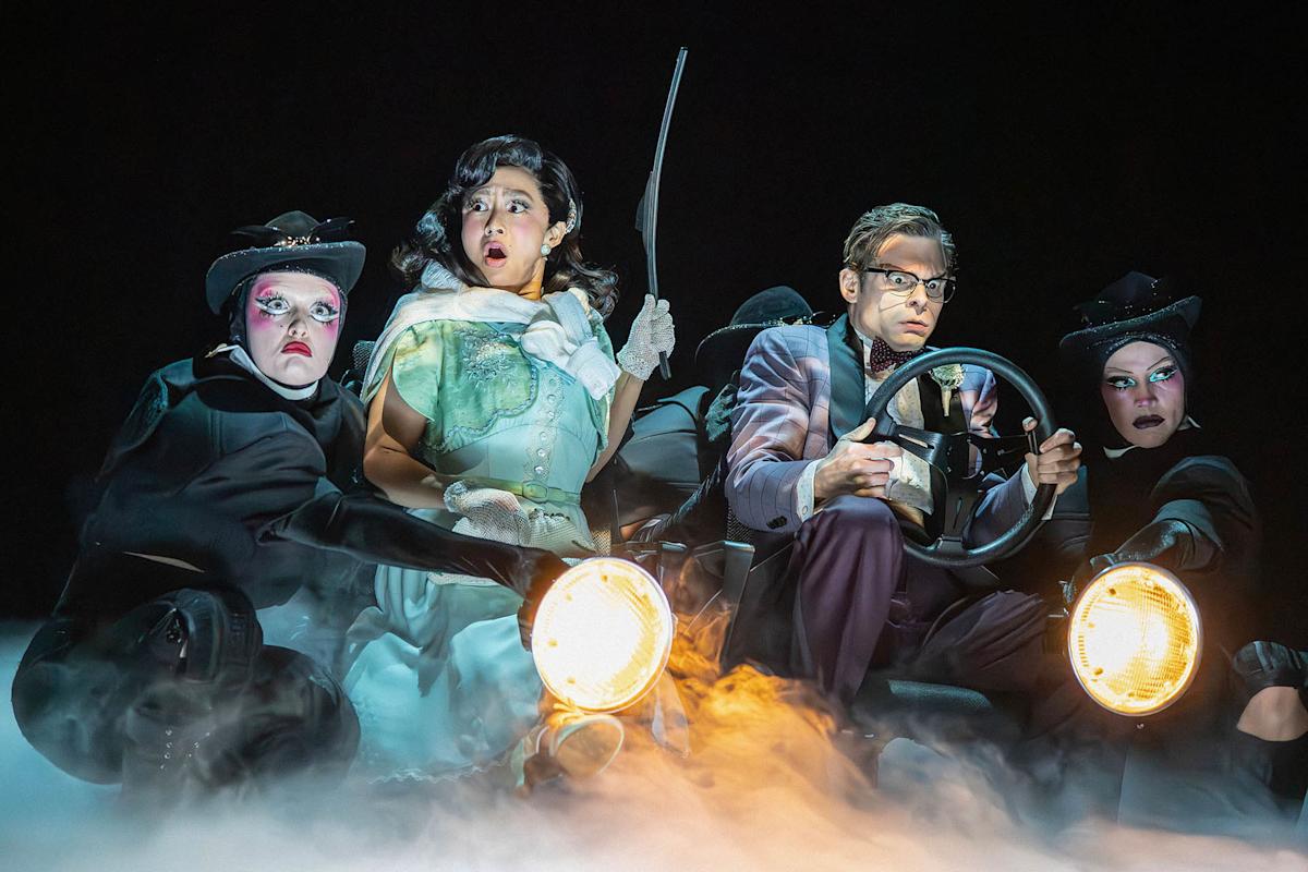 A woman in a green dress and man in glasses drive a car prop, surrounded by three people in black costumes and hats; fog and headlights create a dramatic scene.