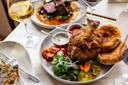 A plate with crispy roast meat on the bone, fresh herbs, sliced cherry tomatoes, pickled vegetables, sauces, and flatbread, surrounded by glasses of wine and additional dishes on a table.