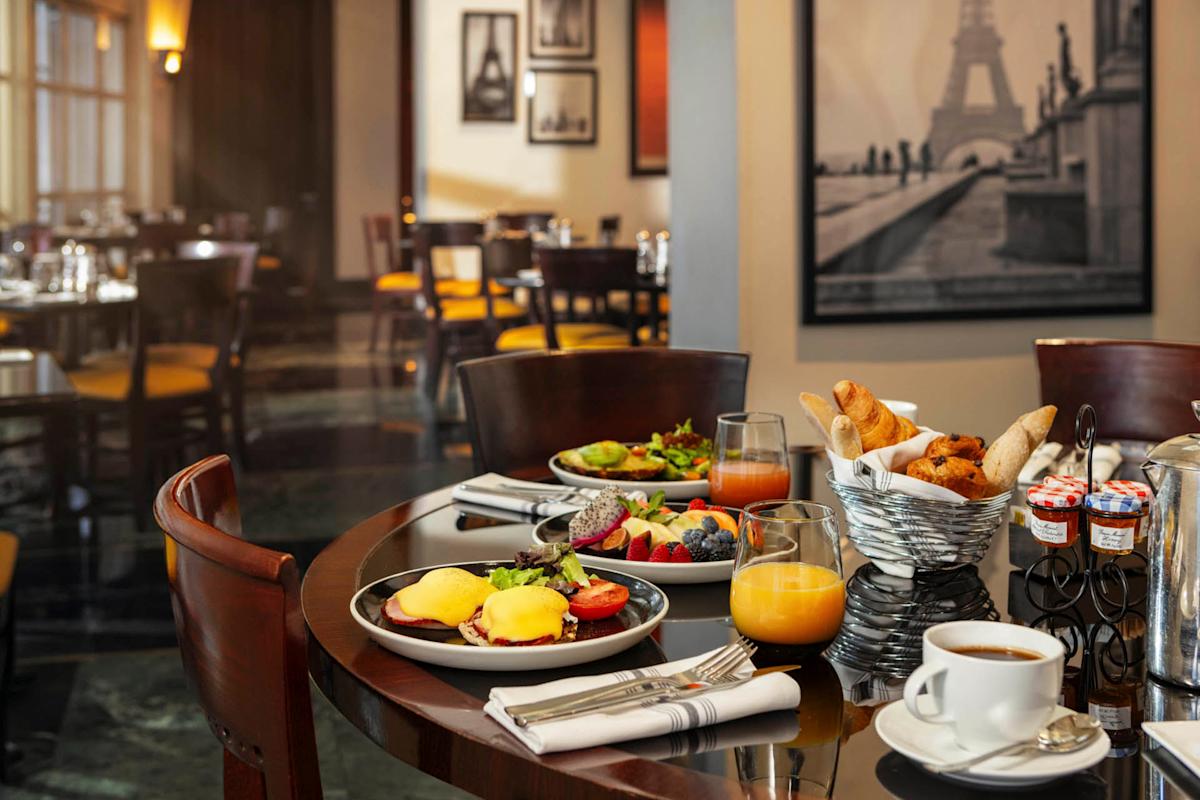 A restaurant table set for breakfast with eggs Benedict, fruit bowl, and croissants in a basket. There's also orange juice and coffee. The background shows a softly lit dining area with framed Paris-themed artwork on the walls.