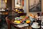 A restaurant table set for breakfast with eggs Benedict, fruit bowl, and croissants in a basket. There's also orange juice and coffee. The background shows a softly lit dining area with framed Paris-themed artwork on the walls.