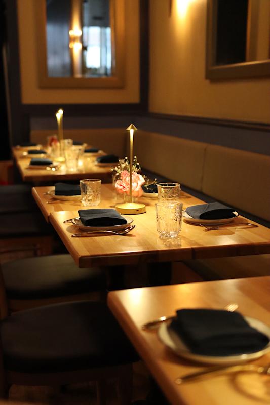 A warmly lit restaurant table set for four, with folded navy napkins, clear glasses, plates, cutlery, a central candle, and a small flower arrangement. The seating is cushioned benches and chairs.