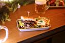 A sandwich filled with sliced meat and vegetables rests on a tray next to a serving of fries garnished with herbs. A mug of frothy beer is in the background, and the table is adorned with greenery.