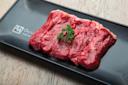 Five slices of raw beef are neatly arranged on a black rectangular plate, garnished with a small sprig of parsley on top. The plate sits on a light wooden surface.