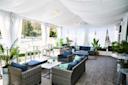 A luxurious outdoor lounge with wicker sofas, blue cushions, small tables, green plants, and white draped curtains, overlooking tennis courts on a sunny day.