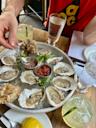 A person spoons sauce onto fresh oysters arranged on a round platter with lemon and garnishes, surrounded by drinks and a partially visible person in a red shirt at a restaurant table.