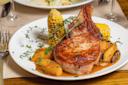 A plate with a grilled pork chop, roasted corn on the cob, carrots, potatoes, and fresh herbs, all topped with a brown sauce, served on a white dish.