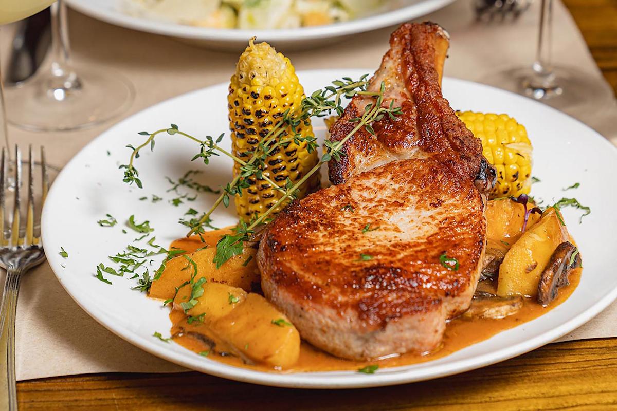A plate with a grilled pork chop, roasted corn on the cob, carrots, potatoes, and fresh herbs, all topped with a brown sauce, served on a white dish.
