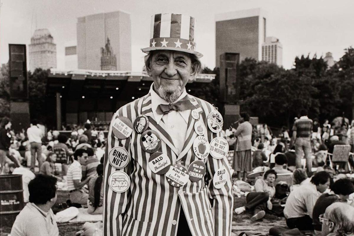 Uncle Sam covered with political buttons stands in a park, holding two “I Love NY” bags. People sit on blankets around him, and tall buildings are visible in the background.