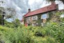 A charming stone house covered in green ivy, with a red metal roof and brick chimneys, surrounded by lush greenery and wildflowers. A lamp post stands nearby, and the sky is cloudy, giving the scene a cozy and serene atmosphere.