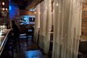 A dimly lit restaurant interior with sheer curtains partially sectioning off seating areas. A person sits at a bar on the left, and there are wooden accents throughout the space. Subtle blue lighting adds ambiance.