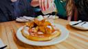 A person pours syrup over a plate of chicken and waffles at a wooden table, with other people and plates visible in the background.