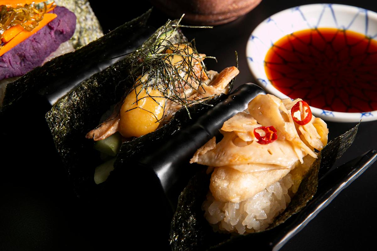 Two hand-rolled sushi pieces with fish, pickled ginger, seaweed, and peppers are displayed next to a small dish of soy sauce on a dark background.
