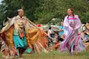 Two women in traditional colorful regalia dance joyfully at an outdoor cultural event. The audience watches, seated on grass, with trees in the background.