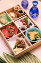 A wooden bento box with assorted Japanese dishes, including sushi, sashimi, vegetables, and side dishes, is arranged neatly on a table with a blue glass sake bottle and cup beside it.