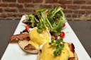 A plate of eggs Benedict topped with hollandaise sauce, garnished with parsley, and served on an English muffin with bacon. A side of fresh mixed greens and cherry tomatoes accompanies the dish. Background shows a brick wall.