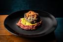 A gourmet dish featuring a stuffed roasted poblano pepper topped with shredded cheese, served on a bed of green sauce and red tomato sauce, presented on a black plate against a dark background.