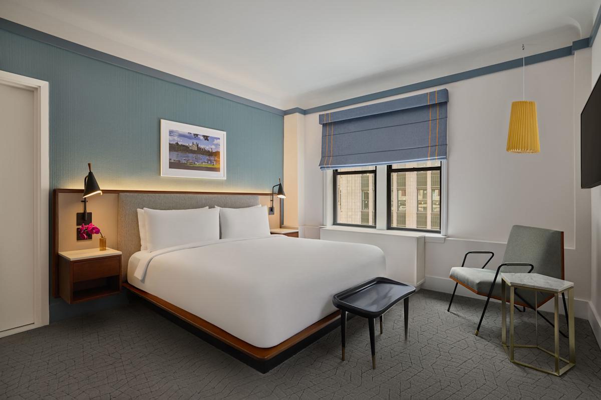 Modern hotel room with a king bed, two nightstands, lamps, wall art, a window with blue blinds, a chair, a small table, and a bench at the foot of the bed. The decor is minimalist and inviting.
