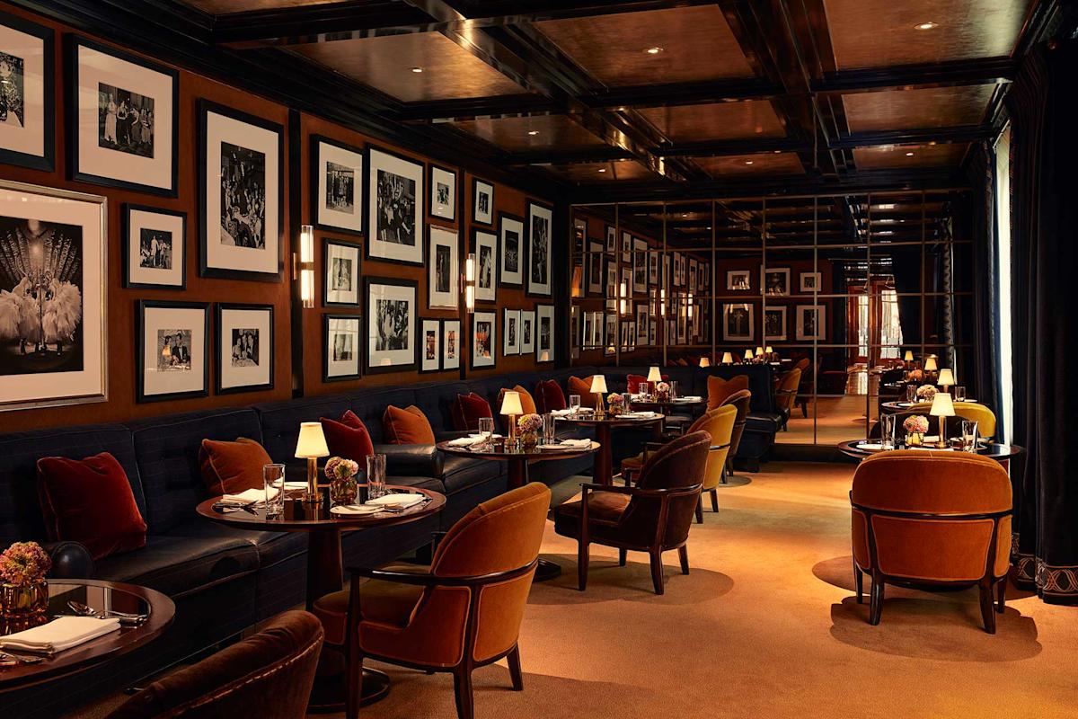 Elegant restaurant interior with dark wood paneling, framed black and white photos on the walls, plush velvet chairs, small round tables set for dining, and warm ambient lighting from table lamps and sconces.