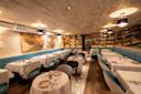 A stylish restaurant interior with light wood ceilings, blue cushioned seating, white tablecloths, and shelves lined with bottles. The warm lighting and abstract wall art create an inviting, upscale ambiance.