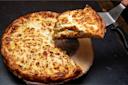 A cheesy, golden brown deep-dish pizza with a slice being lifted out, revealing a thick, gooey cheese interior. The pizza is on a wooden board, set against a dark background.