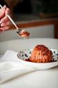A hand holding a spoon hovers over a dessert topped with crumbled bits and drizzled with caramel sauce, served in a decorative bowl on a white napkin.