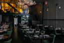 Dimly lit modern restaurant with green chairs, tables set with plates and glasses, a floral chandelier, exposed brick wall, and an illuminated wine rack on a mezzanine level.
