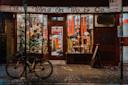 A bicycle is parked outside Wing On Wo & Co., a brightly lit shop with Chinese decorations, red lanterns, and festive banners. The sidewalk is scattered with confetti, and the shop windows display various items.