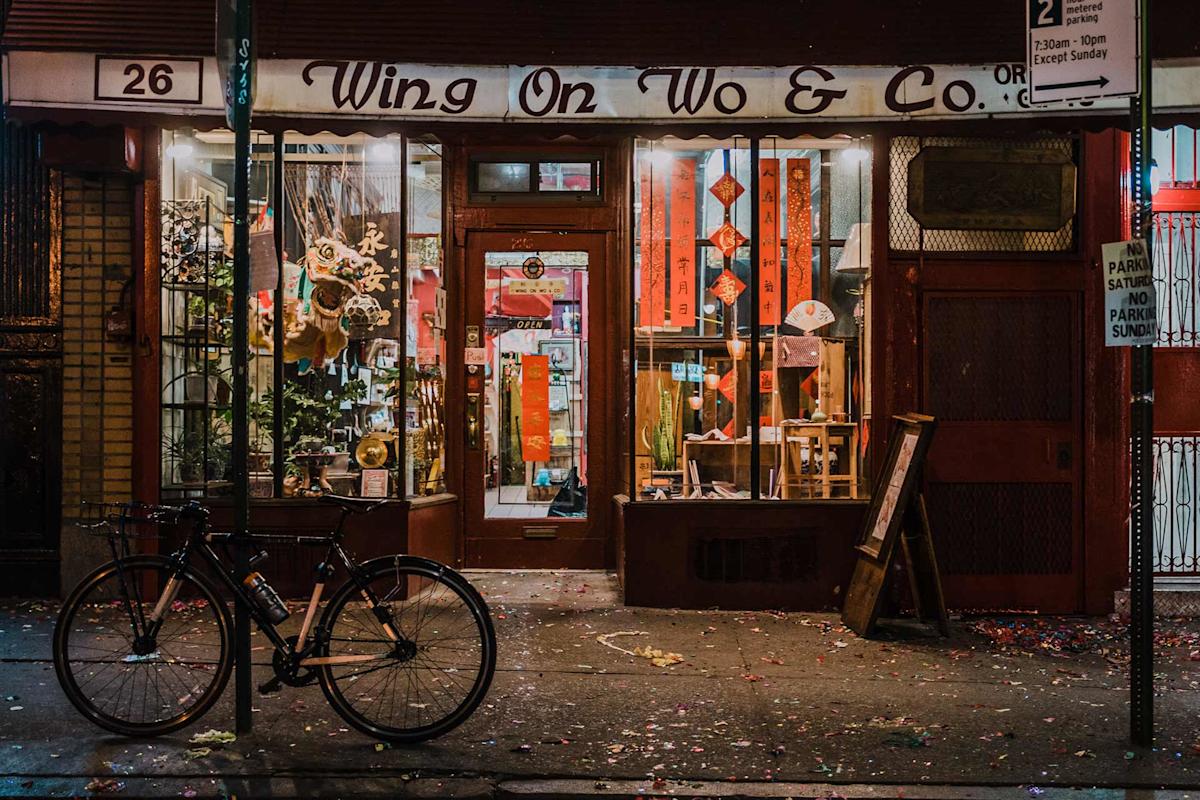 A bicycle is parked outside Wing On Wo & Co., a brightly lit shop with Chinese decorations, red lanterns, and festive banners. The sidewalk is scattered with confetti, and the shop windows display various items.