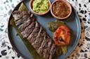 A plate with grilled steak on a banana leaf, served with guacamole, salsa, a grilled jalapeño, grilled tomato, and cactus on a terrazzo surface.