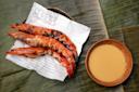 Grilled shrimp served on a piece of printed paper next to a bowl of sauce, placed on a banana leaf.
