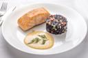 A plate with a piece of cooked salmon, a round serving of black rice mixed with diced vegetables, and a portion of creamy yellow sauce garnished with dill, all arranged neatly on a white dish.