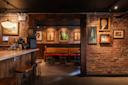 A cozy, dimly lit bar with exposed brick walls. Wooden stools line the counter on the left, while the seating area in the back features a wooden bench and chairs. Various framed portraits and artwork adorn the walls.
