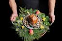 A person holds a round tray decorated with pine branches, leaves, tulips, and yellow flowers, featuring a baked pastry dome placed on a bed of smooth stones.