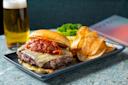 A cheeseburger with sauce sits on a rectangular plate next to a pile of potato chips. A tall glass of beer and a menu are in the background on a marble table.