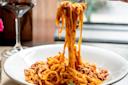A plate of tagliatelle pasta is topped with a rich, red tomato sauce. A fork twirls a portion of the pasta above the plate. A glass of red wine sits nearby on the table.