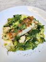 A fillet of seared fish sits atop a bed of greens, beans, fresh herbs, and corn, served on a white plate with a light sauce, garnished with dill and mint.