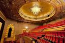Opulent theater interior with red velvet seats, ornate gold ceiling featuring a large chandelier, and decorative architectural details along the walls and balcony.