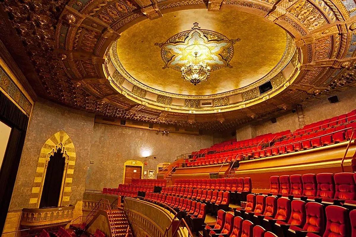 Opulent theater interior with red velvet seats, ornate gold ceiling featuring a large chandelier, and decorative architectural details along the walls and balcony.