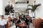 A bustling cafe with large windows and lush indoor plants. People are seated at tables, engaged in conversation, while a server attends to guests. Natural light fills the space, creating a warm and inviting atmosphere.