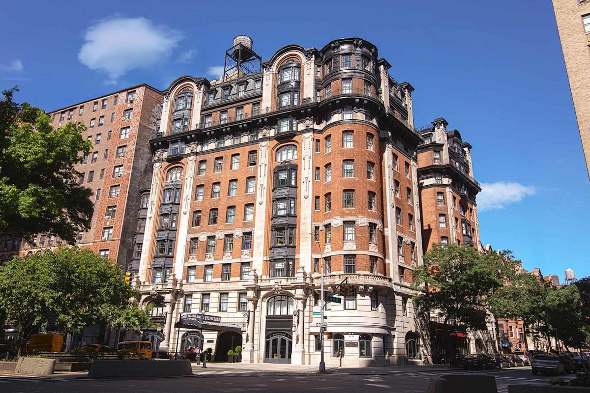 A historic, multi-story brick building with ornate architectural details stands on a sunny street corner, surrounded by trees and other buildings under a bright blue sky with a few clouds.