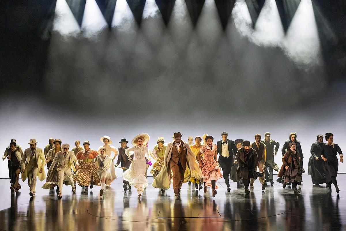 A large group of people in period costumes run towards the camera on a stage, illuminated by bright spotlights against a misty background.