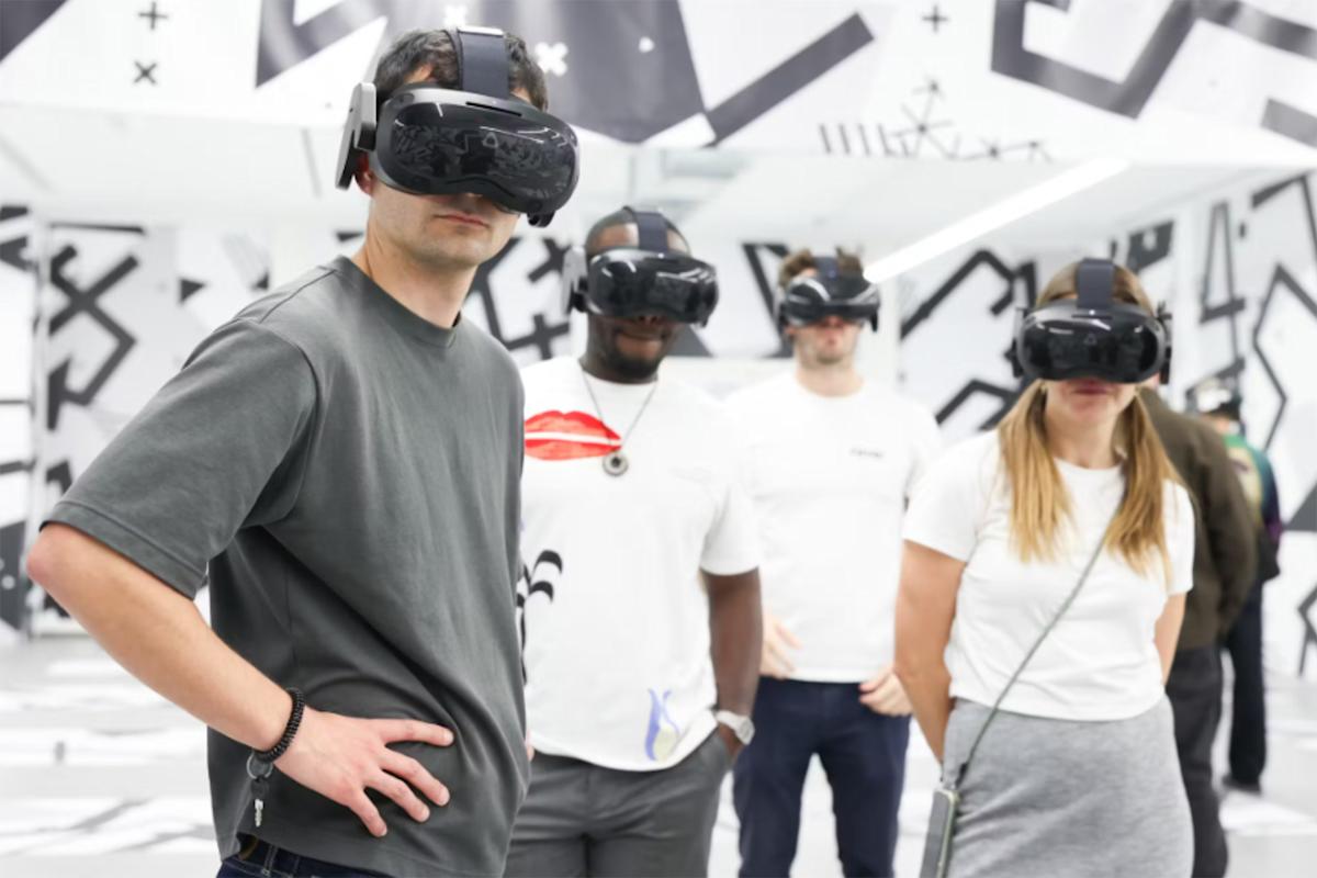 A person wearing a virtual reality headset stands with arms raised in front of a black-and-white geometric mural. The person wears a patterned sweater and black pants, appearing to interact with the VR environment.