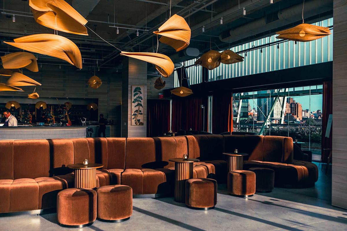 Modern lounge with brown velvet seating, small round tables, unique leaf-shaped ceiling lights, large windows, and a view of a cityscape in the background. Warm sunlight casts shadows across the stylish interior.